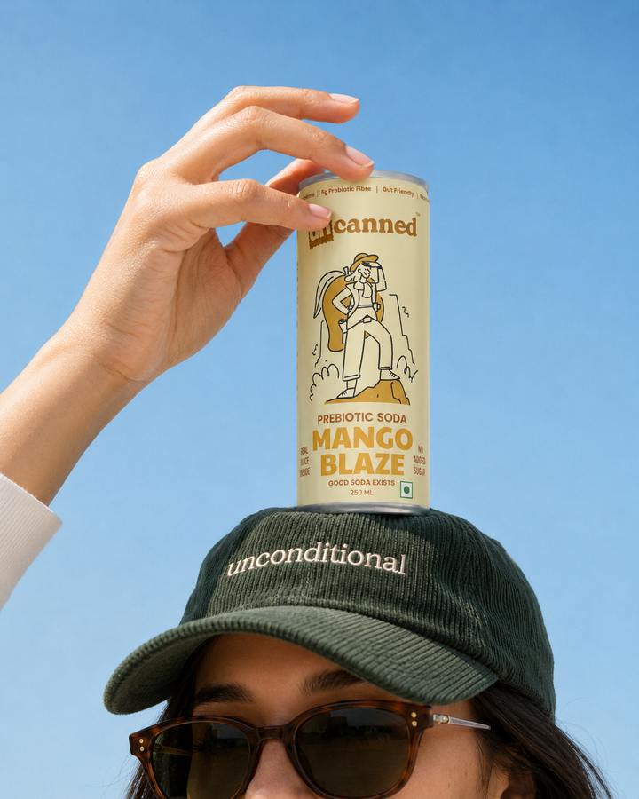 A person outdoors against a clear blue sky holds a can of Uncanned Prebiotic Sock Mango Blaze on top of their green corduroy cap.